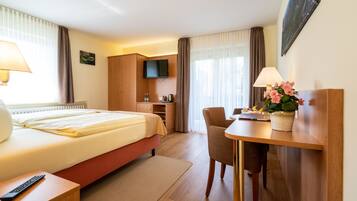 A hotel room with a bed, bedside table, TV, desk with chair, and a small kitchenette area.