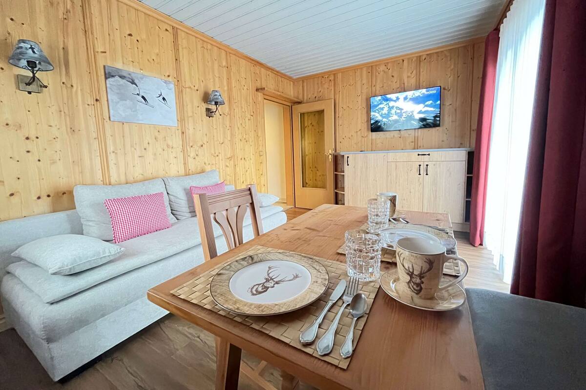 Ett mysigt rum med träinredning, en soffa med kuddar, ett matbord dukat med tallrikar, bestick och glas, och en tv som är monterad på väggen.