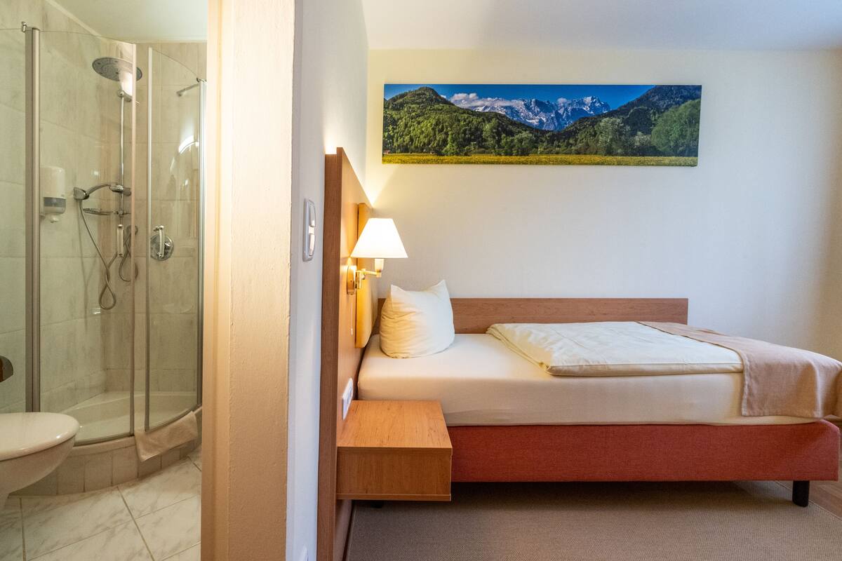 Ett hotellrum med en säng, ett sängbord, en lampa, en dusch och en tavla på väggen.