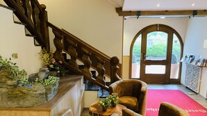A reception area with a wooden staircase, a reception desk, two armchairs, and a red carpet leading to an arched doorway. - Hotel Garni Brunnthaler (Garmisch-Partenkirchen)