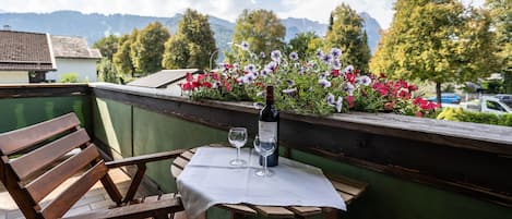 A wooden table with a white tablecloth, two wine glasses, and a bottle of wine on a balcony with a view of mountains and trees.
