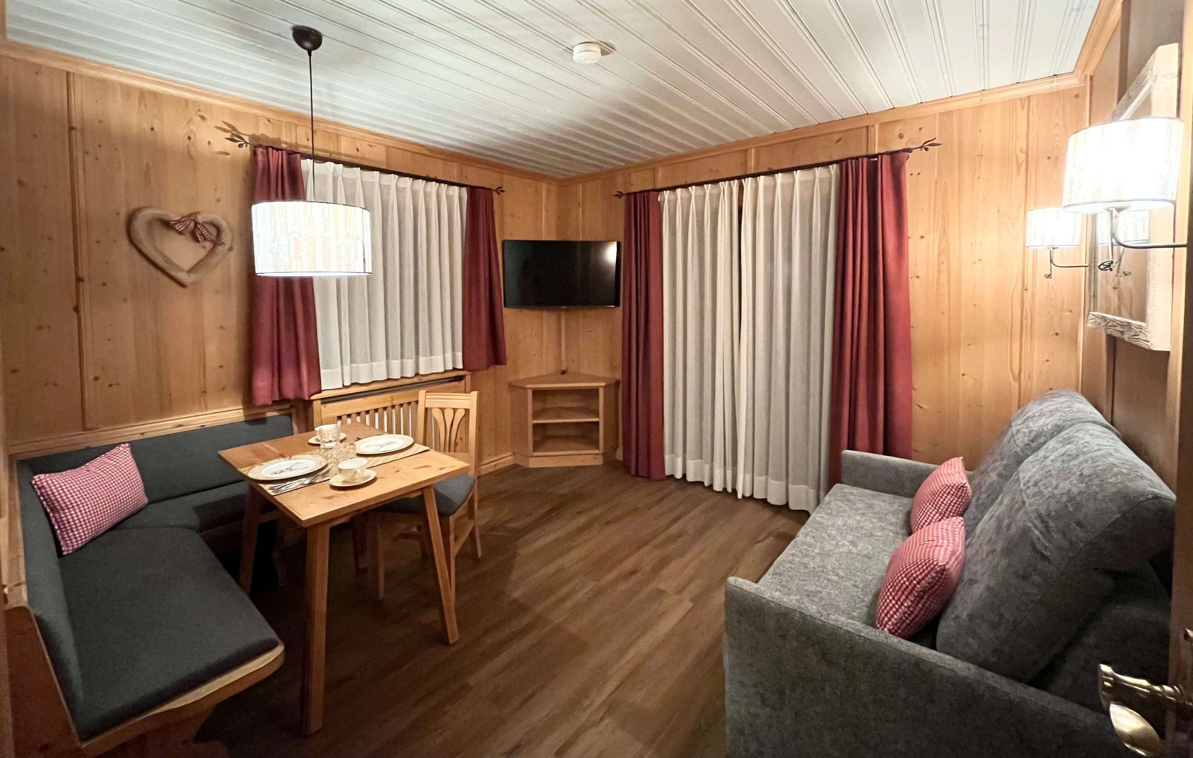 A cozy room with a wooden interior, a dining table with chairs, a sofa, and a television mounted on the wall.