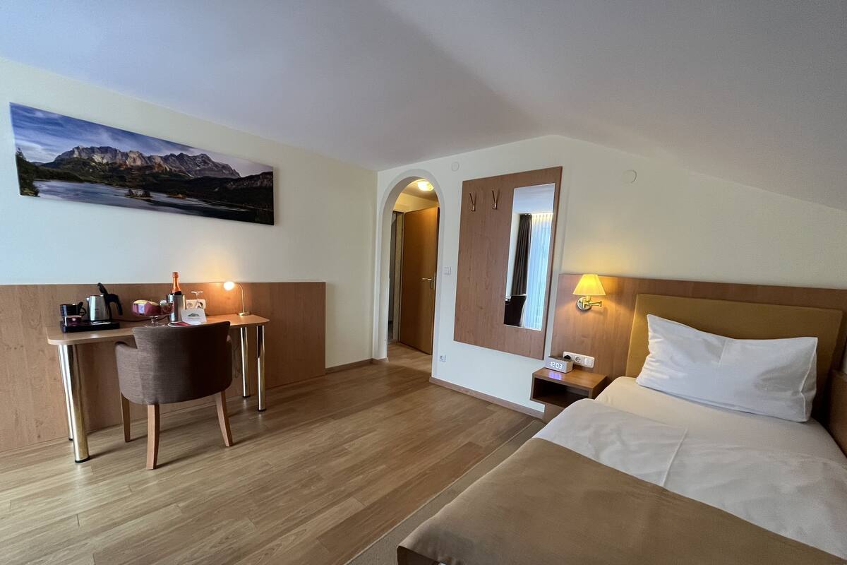 Ett hotellrum med en säng, ett skrivbord med en stol, ett litet bord med en lampa, en spegel och en tavla på väggen.