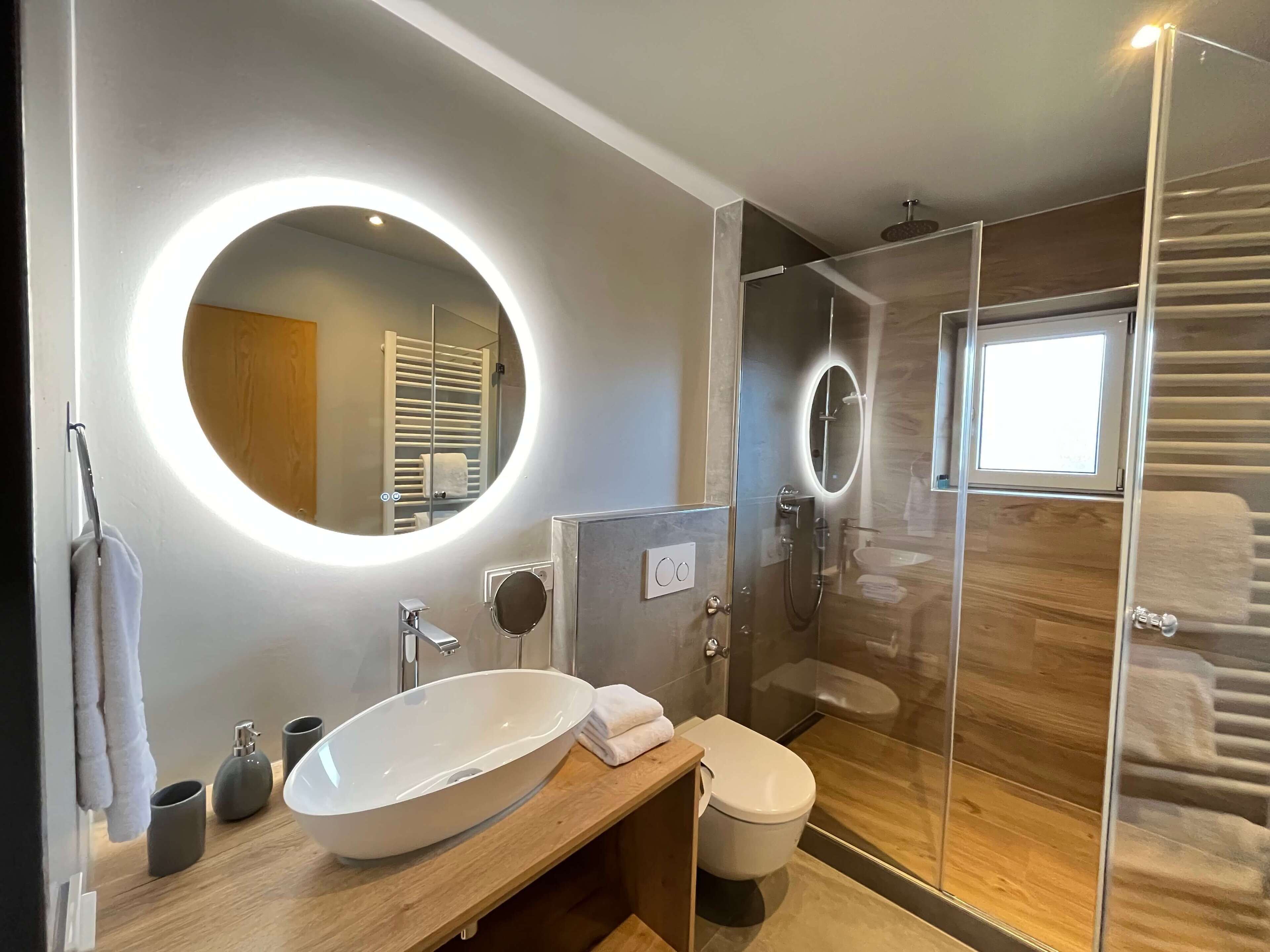 A modern bathroom with a large oval sink, a round mirror, a glass-enclosed shower, and a toilet.