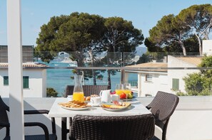Un balcón con muebles de mimbre, alimentos para el desayuno y vistas al mar y a los árboles.