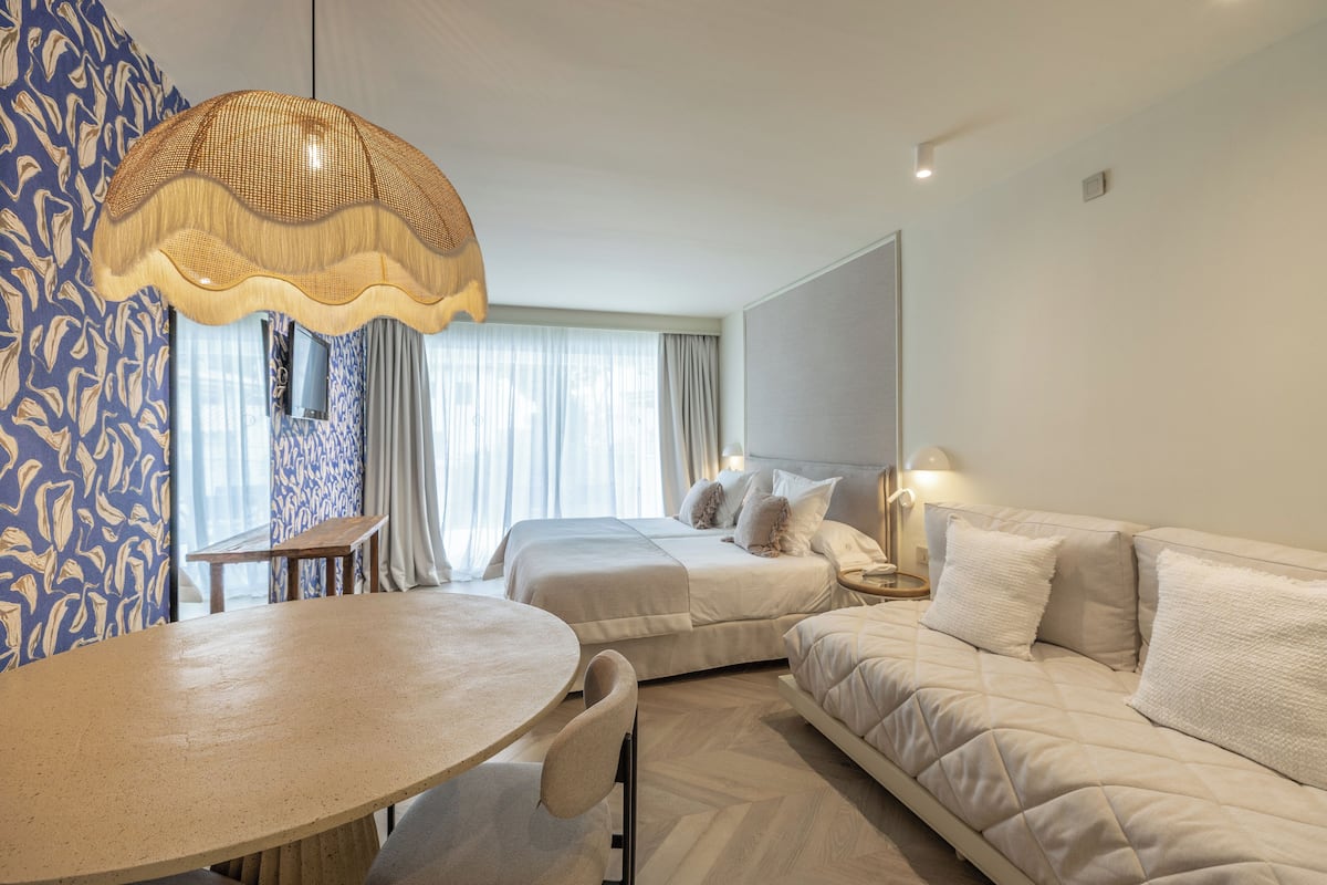 Ein Hotelzimmer mit einem runden Tisch, zwei Betten, einem Sofa, einer großen Pendelleuchte und Blick auf die Stadt durch das Fenster.