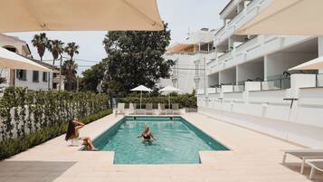 Zona de piscina con sillones reclinables y sombrillas, rodeada de edificios y vegetación.