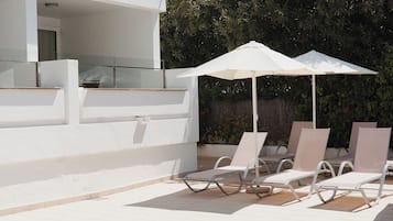 Uma área de estar moderna ao ar livre, com cadeiras de lounge brancas e guarda-sóis, um edifício branco com varanda de vidro e um fundo de folhagem densa.