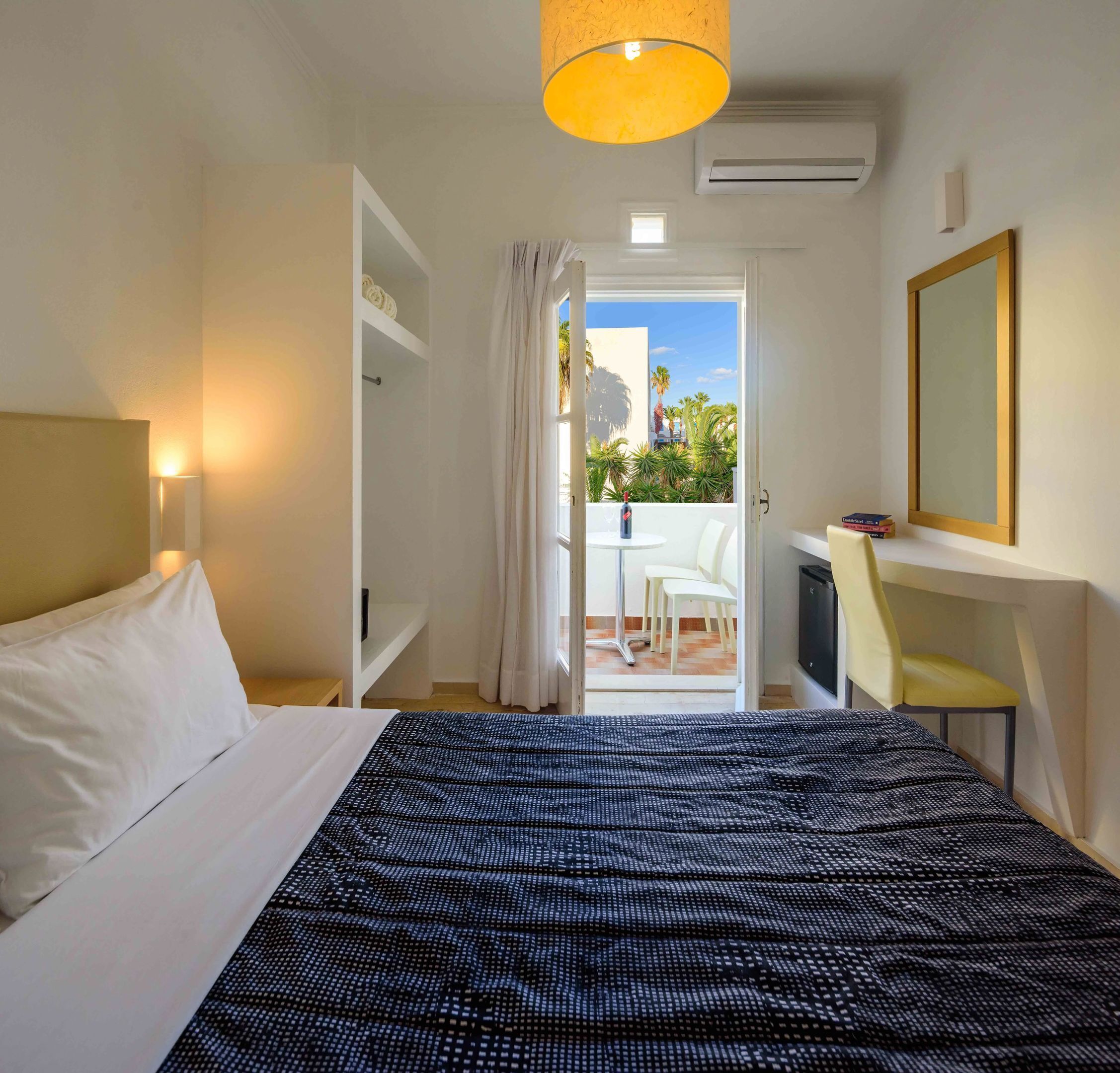 Een hotelkamer met een bed, een bureau, een stoel en een balkon met planten.