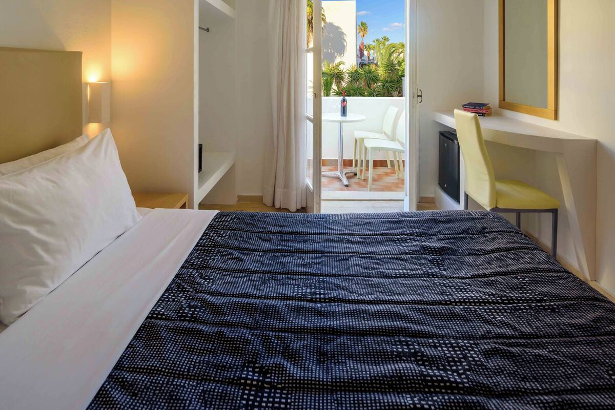 Une chambre d’hôtel avec un lit, un bureau, une chaise et un balcon agrémenté de plantes.