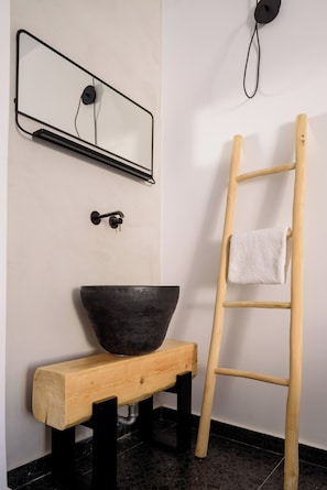 A bathroom with a wooden bench, a black basin, a towel on a wooden ladder, a wall-mounted mirror, and a towel rack.