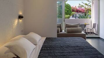 A modern bedroom with a large bed, a balcony with seating, and a view of greenery and buildings.