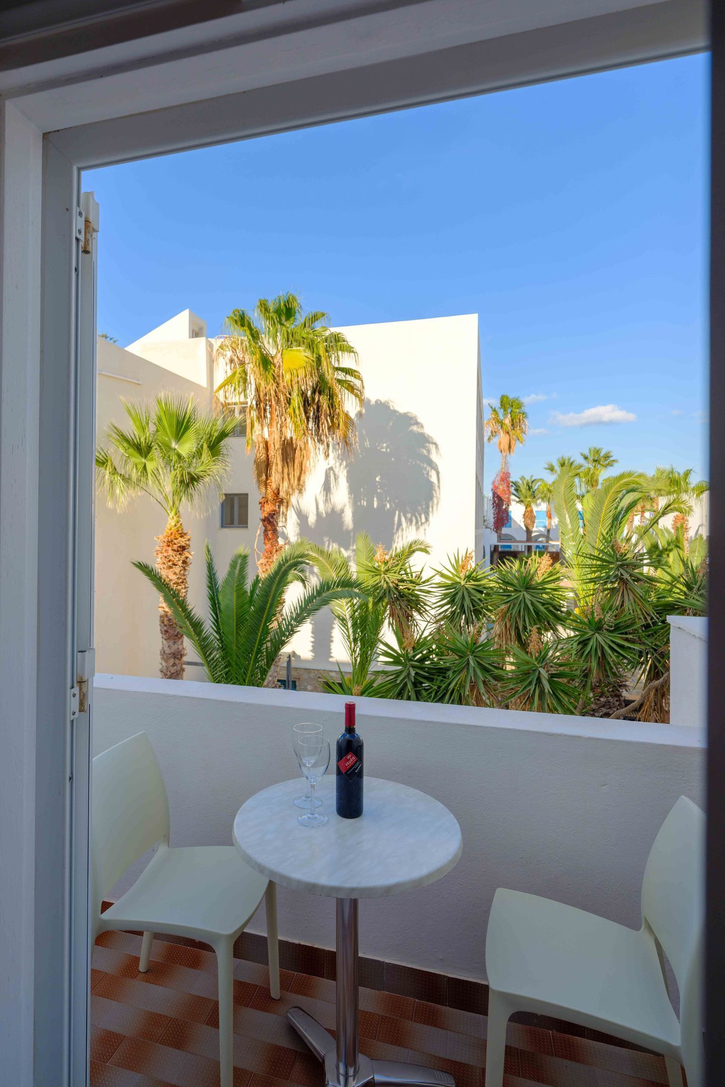 A balcony with a table and chairs, a bottle of wine, and a view of palm trees and buildings.