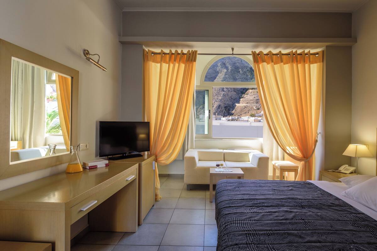 Une chambre d’hôtel avec un lit, un bureau avec une télévision, un canapé et une vue sur un paysage montagneux par la fenêtre.