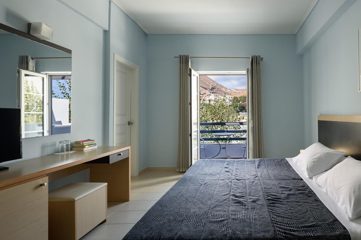 Un dormitorio con cama, escritorio, televisor y vistas a un balcón con una montaña al fondo.