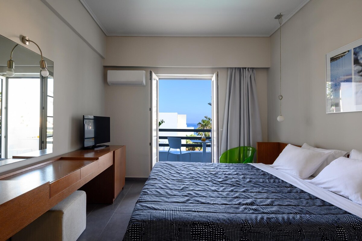 Habitación de hotel con cama, escritorio, televisor, balcón con vista al mar y un cuadro en la pared.
