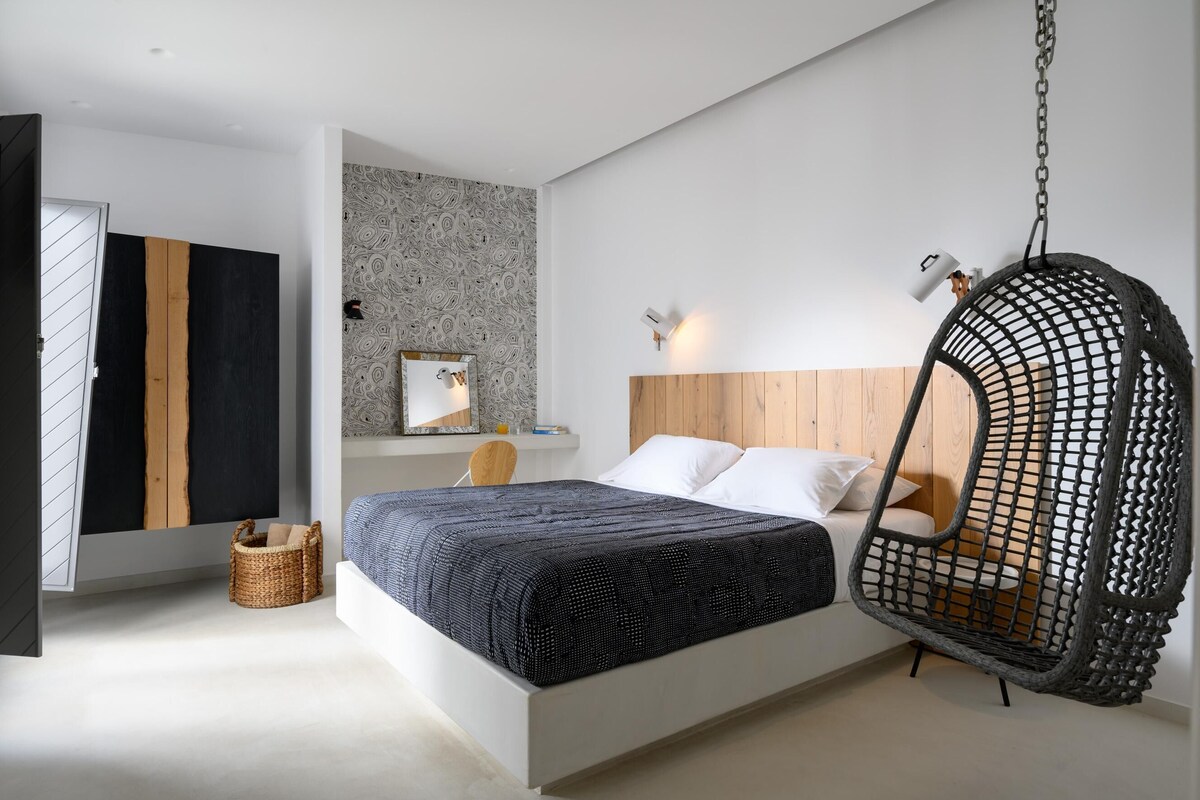 Un dormitorio moderno con cama, una silla colgante, cabecero de madera y televisor montado en la pared.