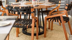 Un comedor con una mesa redonda de madera dispuesta para seis personas, rodeada de sillas negras y de madera. Un lámpara tejida cuelga sobre la mesa, y hay una pared de madera con plantas verdes al fondo.