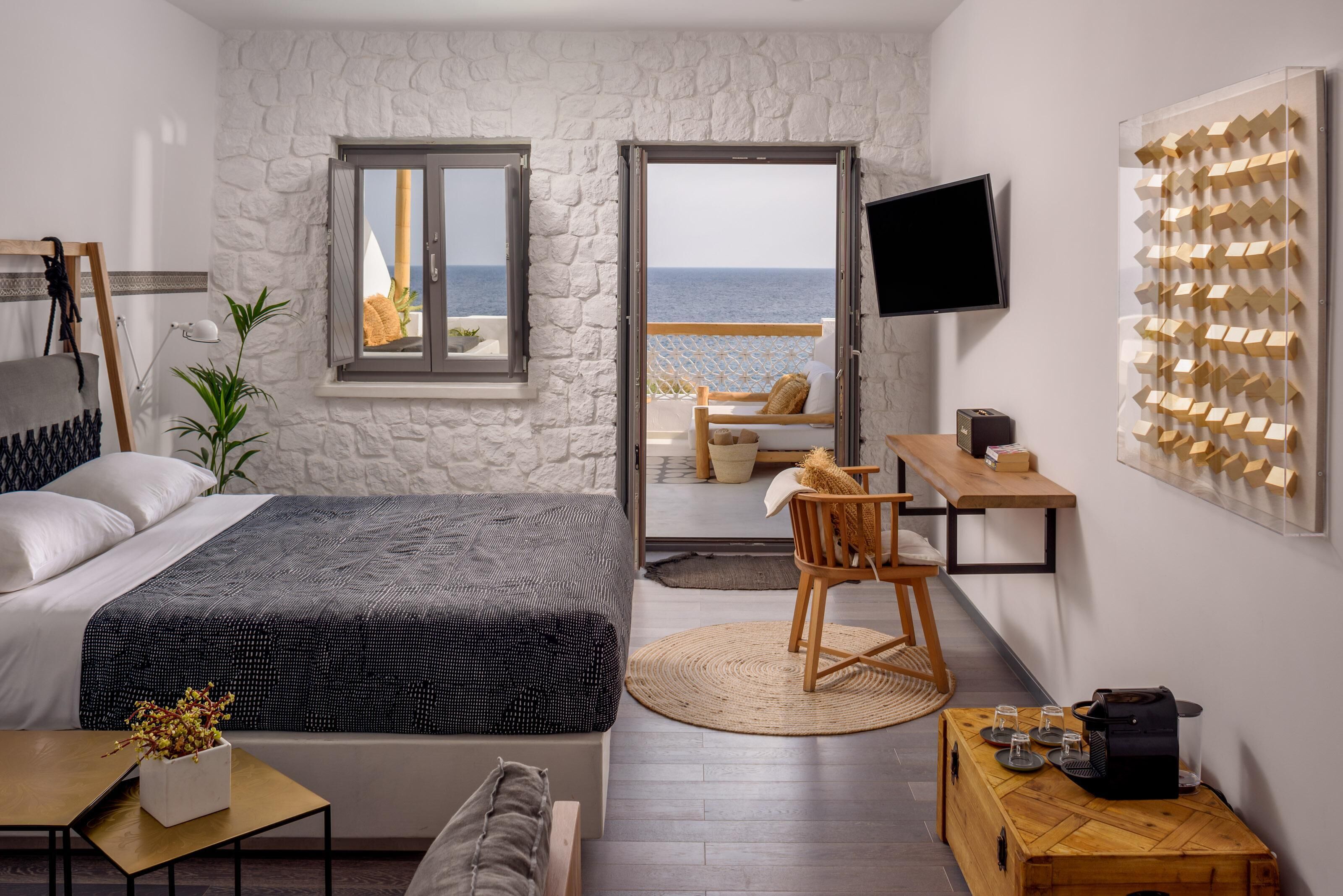 A modern bedroom with a bed, a desk, a chair, and a view of the sea through an open door.