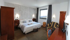 A hotel room with a bed, bedside tables, a sofa, a window with curtains, and a view of the outdoors.
