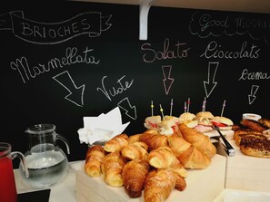 A breakfast buffet with croissants, sandwiches, and a chalkboard menu listing items like brioches, marmellata, and eclima.
