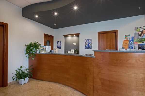 A reception area with a wooden reception desk, potted plants, framed artwork, and a ceiling with recessed lighting.