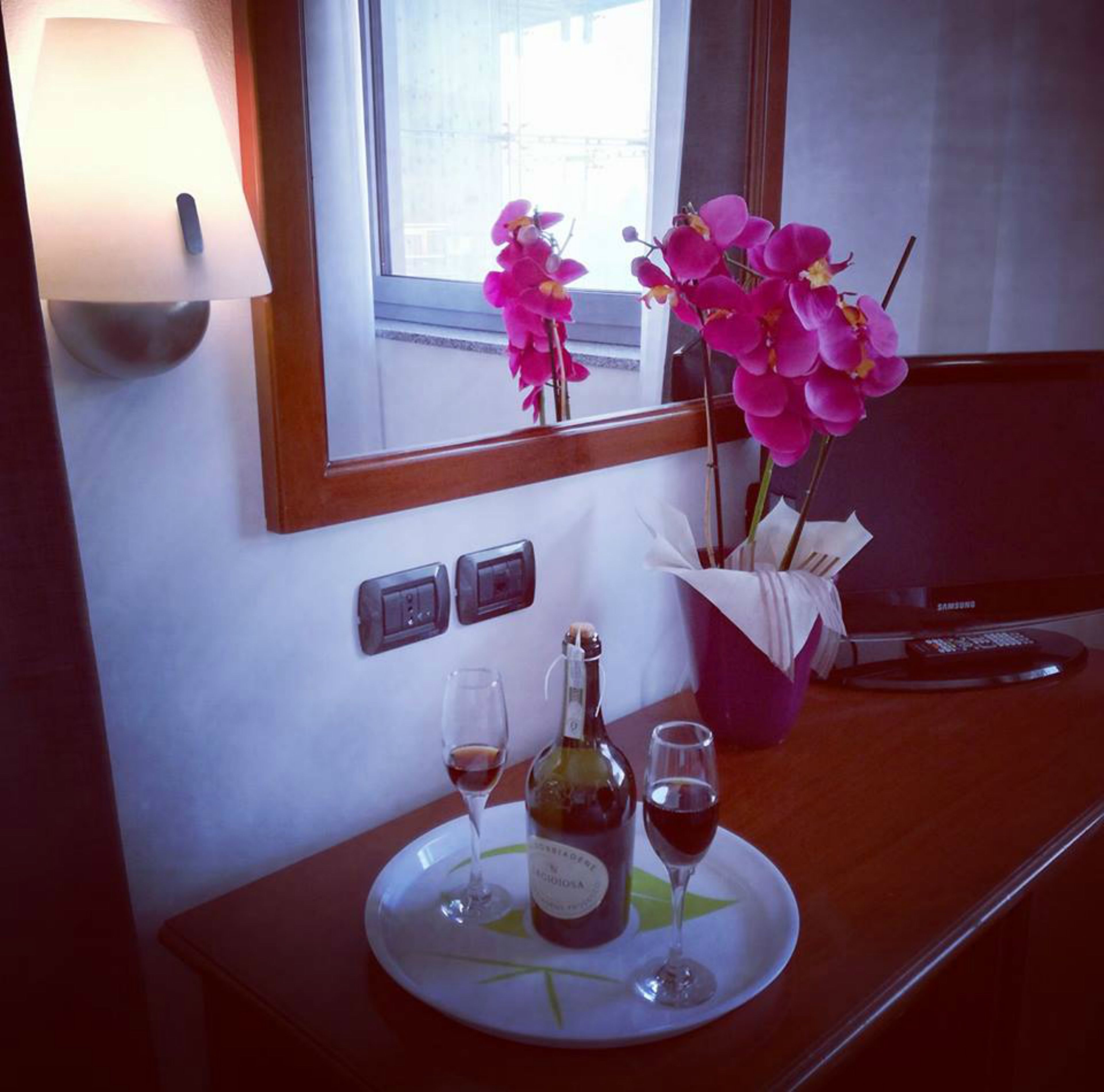Una camera d'albergo con una scrivania in legno, uno specchio, un vaso con orchidee rosa, una bottiglia di vino e due bicchieri da vino.