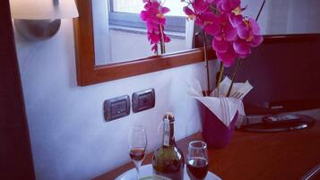 Una camera d'albergo con una scrivania in legno, uno specchio, un vaso con orchidee rosa, una bottiglia di vino e due bicchieri da vino.