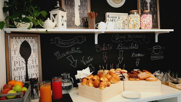 A breakfast buffet with various pastries, a chalkboard menu, and a clock on the wall. - Medea (Alba)