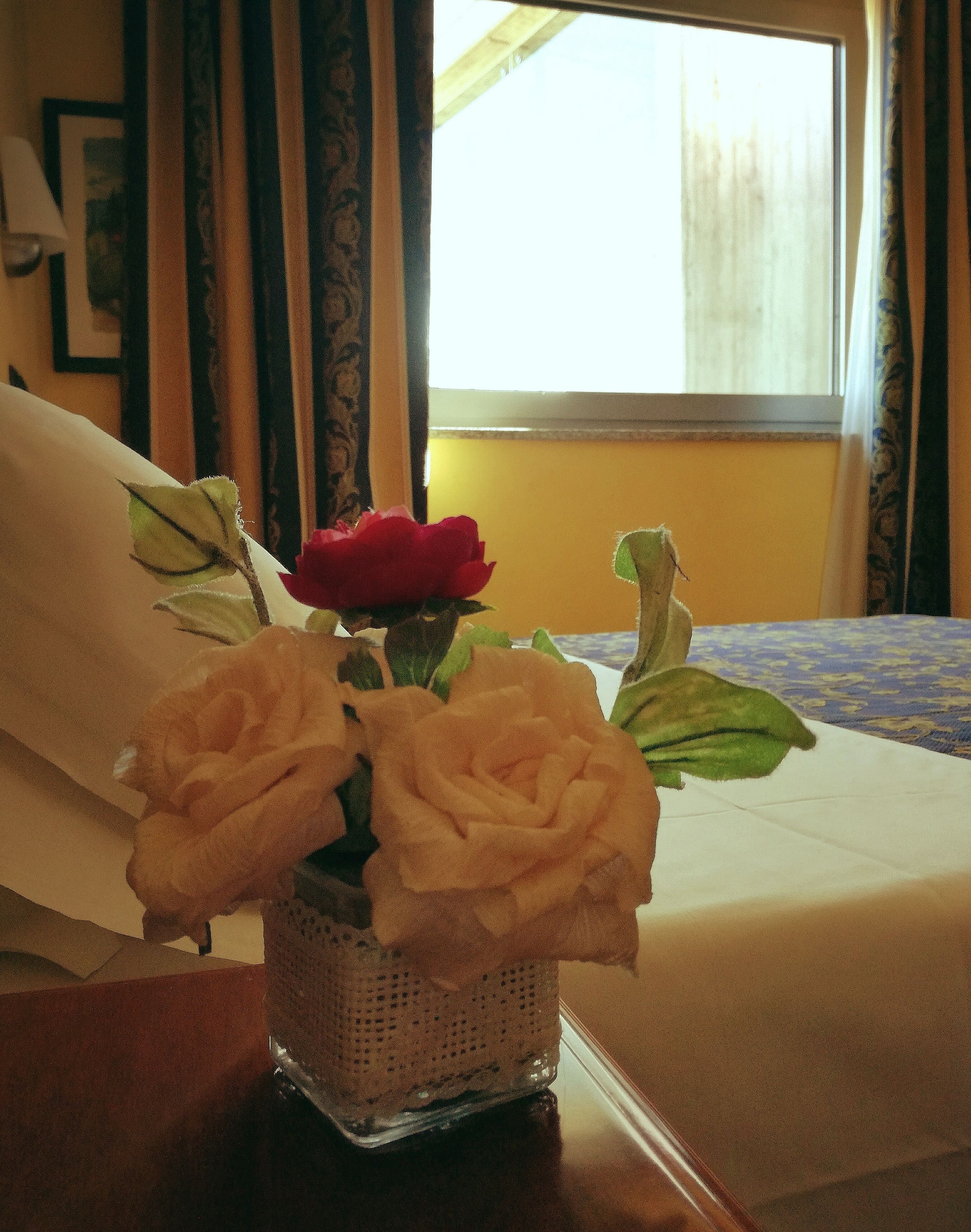 Ein kleiner Glasvase mit zwei weißen Rosen und einer roten Rose auf einem Holztisch in einem Hotelzimmer.