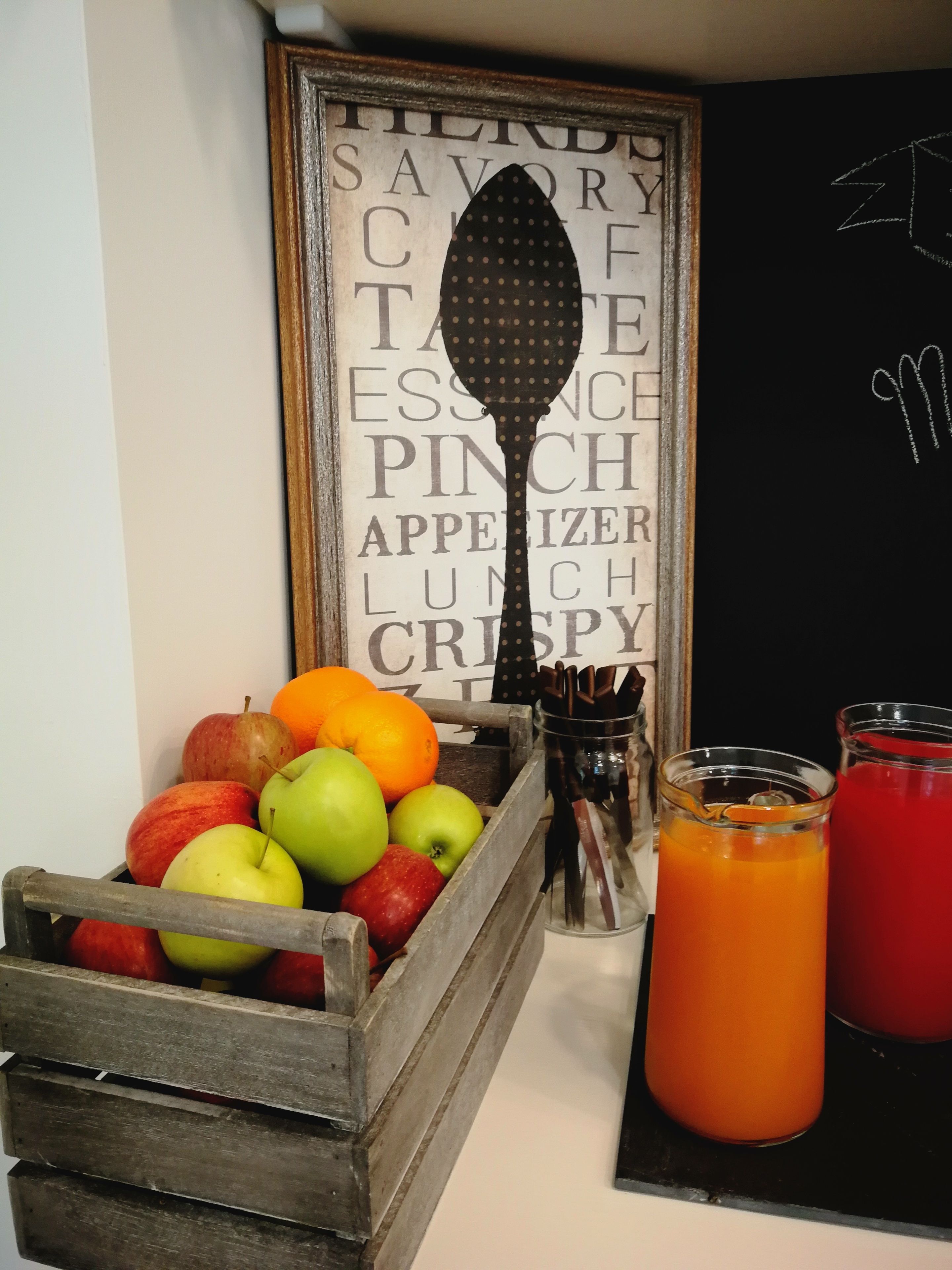 A wooden crate filled with apples and oranges, a glass pitcher with a drink, and a framed word cloud on the wall.