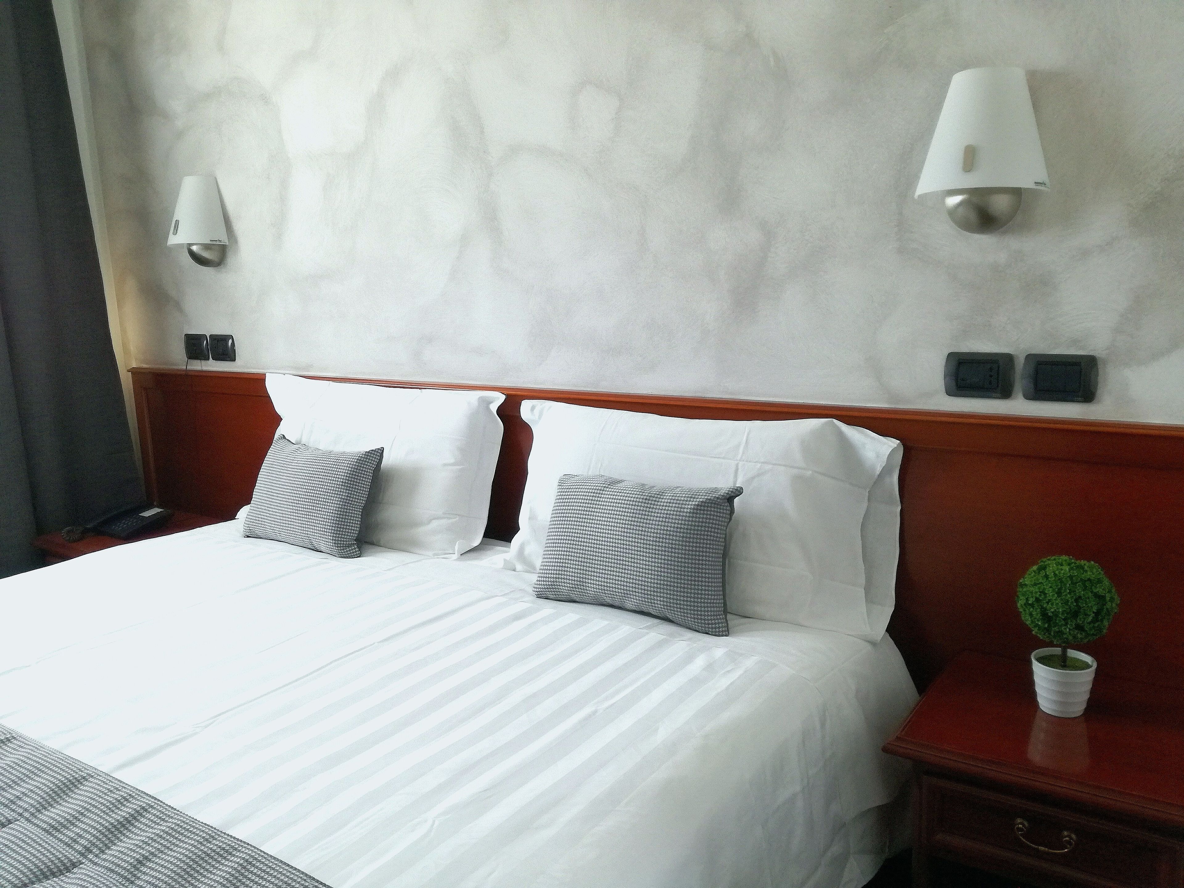 A hotel room with a large bed, two bedside tables, a nightstand, a small potted plant, and two wall-mounted lamps.