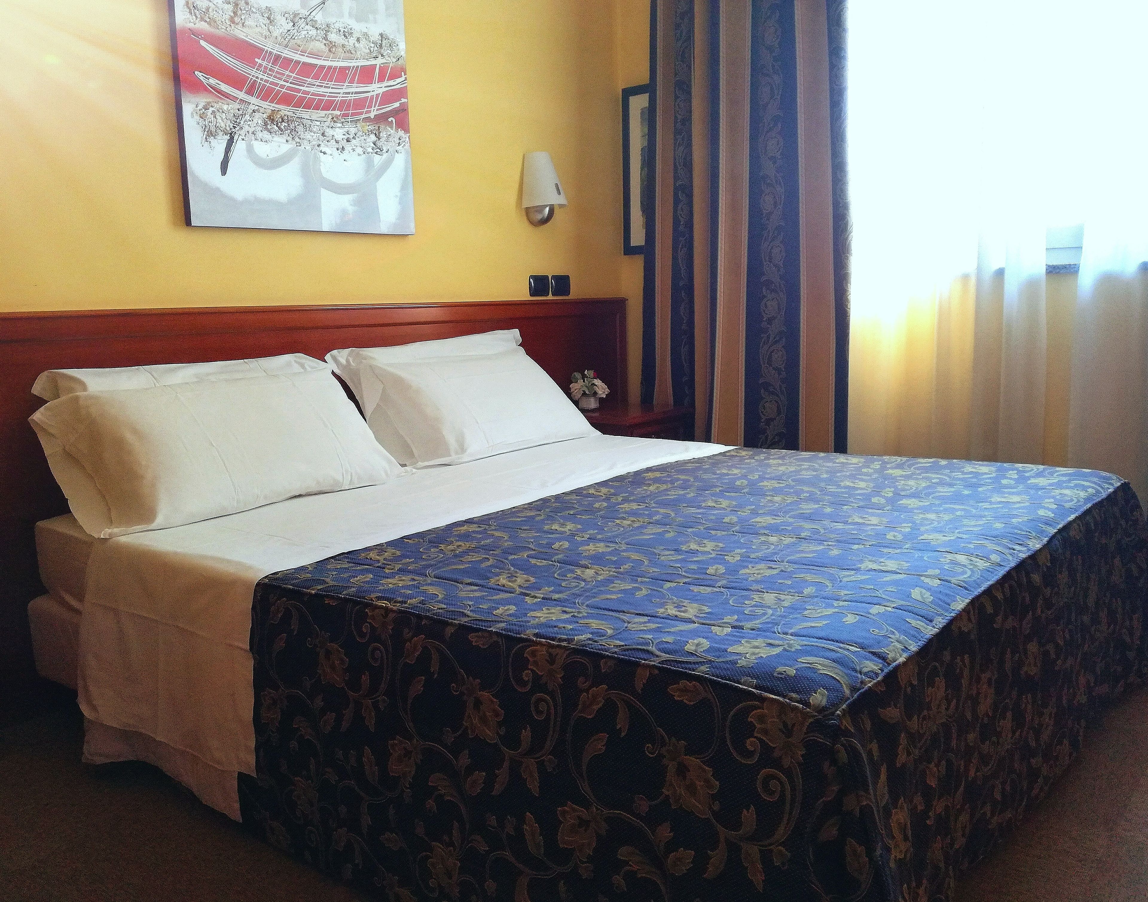 A hotel room with a bed, a headboard, a wall-mounted light, a painting, and a window with curtains.