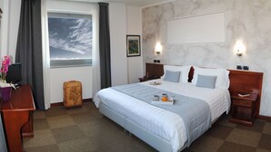 A hotel room with a large bed, a desk with a computer, a suitcase, and a window with curtains. - Medea (Alba)