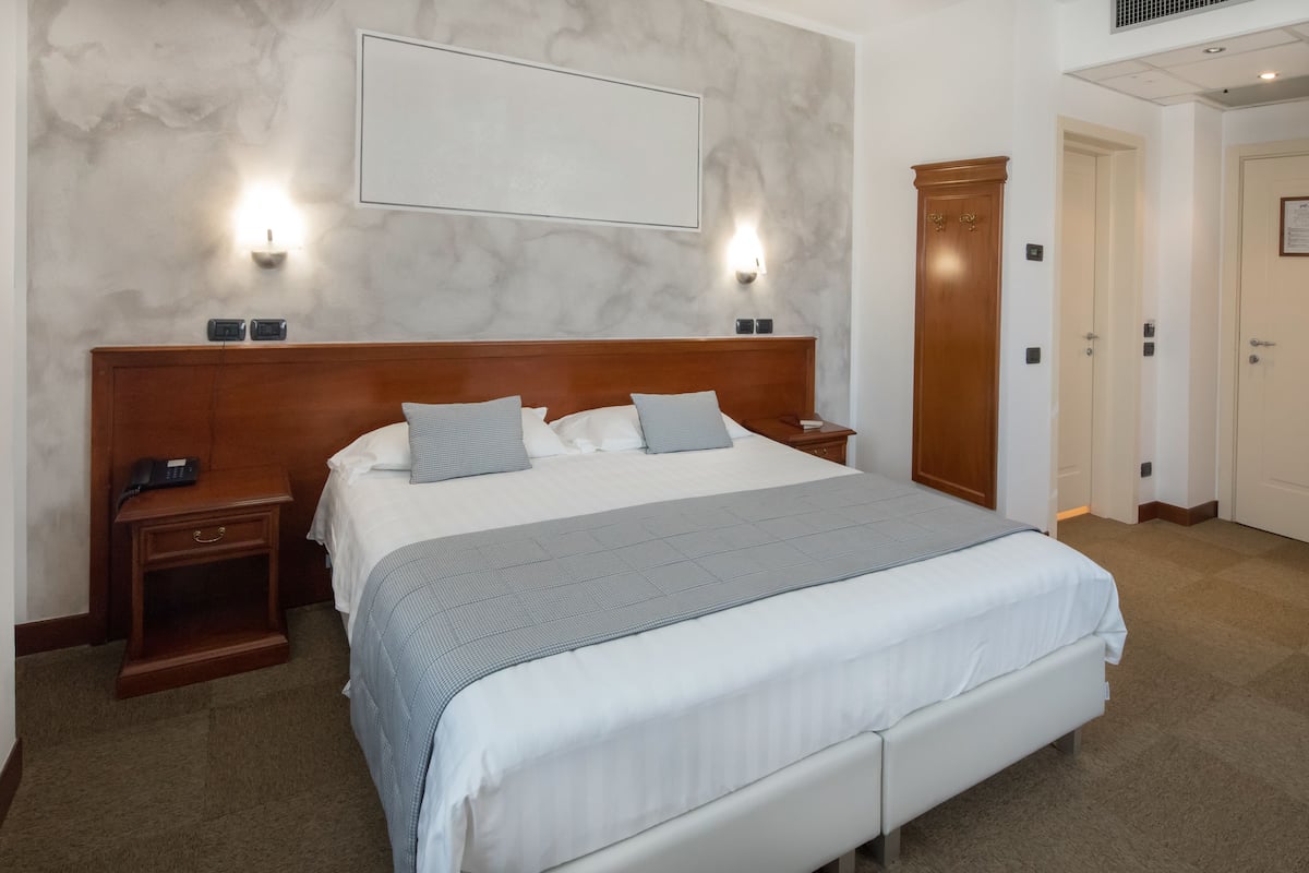 Habitación de hotel con una cama grande, cabecero de madera, dos mesitas de noche, un armario y una lámpara de pared.