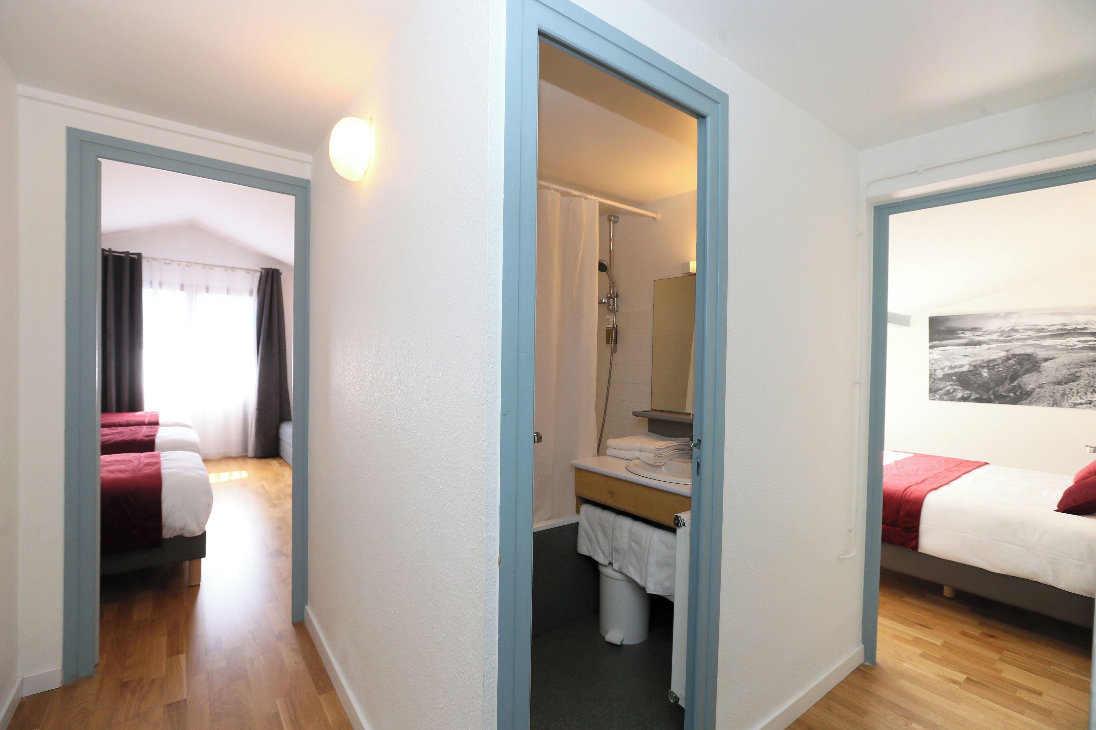 Habitación de hotel con piso de madera, una cama con cubrecama rojo y blanco, baño con ducha y un ventanal con vistas.