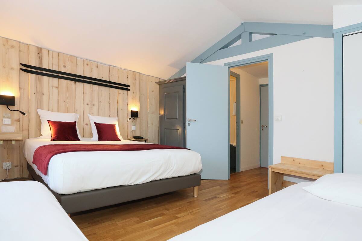 A modern bedroom with a wooden headboard, a bed with white and red bedding, a bedside table with a lamp, and a wooden bench.