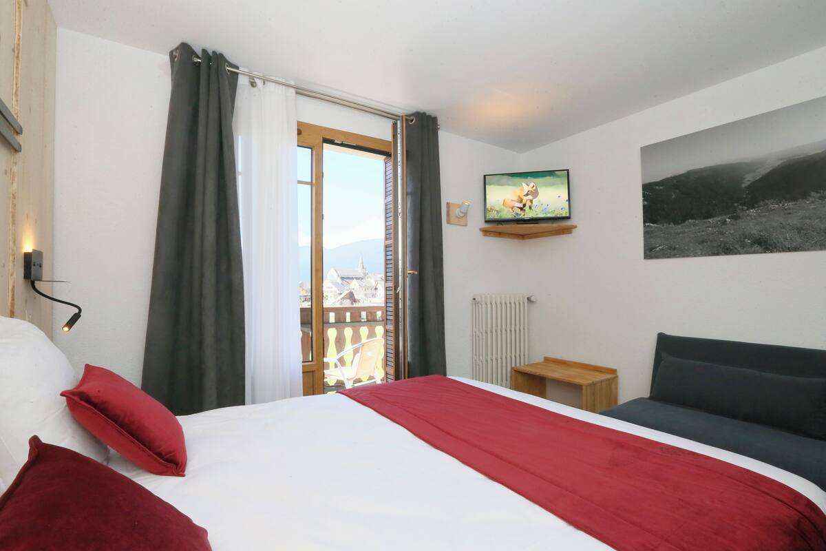 A hotel room with a bed, a TV, a sofa, a window with curtains, and a view of the outside.