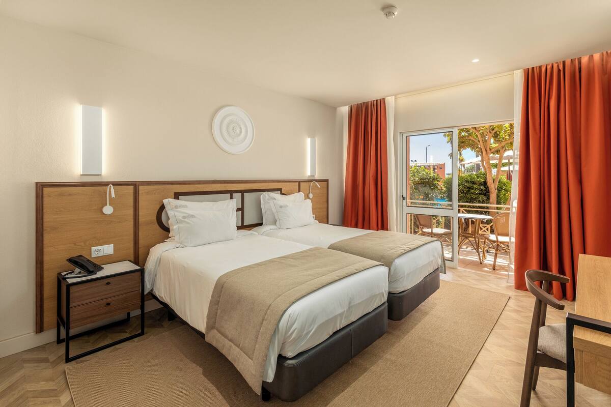 Une chambre d’hôtel avec deux lits, un bureau avec une chaise, une petite table, une table de chevet et une fenêtre avec des rideaux.