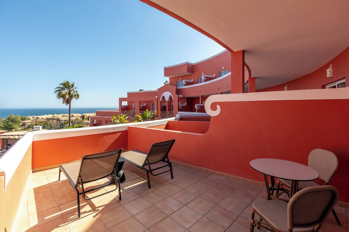 Un balcon avec vue sur la mer, un palmier et un bâtiment aux murs rouges et blancs.