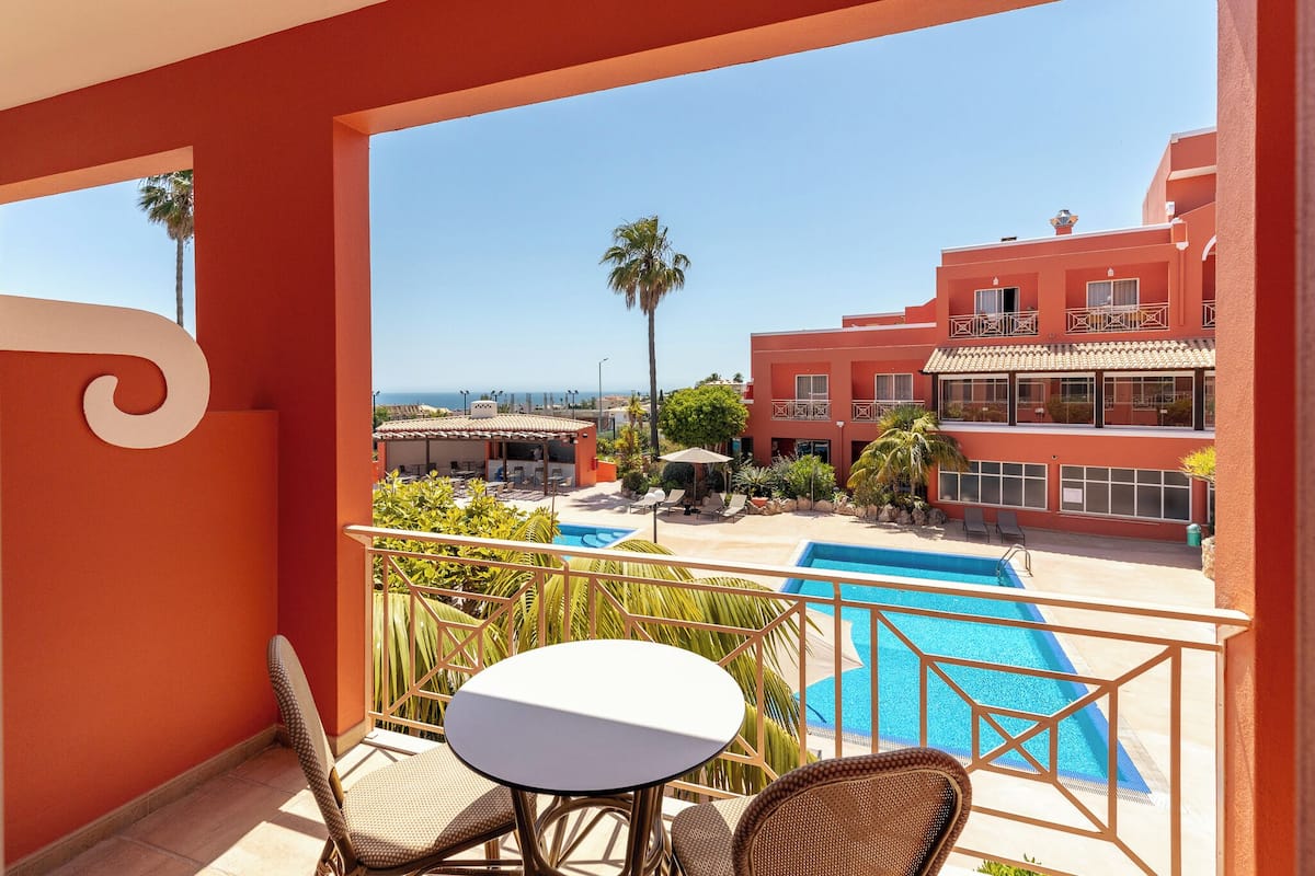 Un balcon avec une piscine, des fauteuils en osier et une vue sur un bâtiment rouge avec un toit en tuiles.