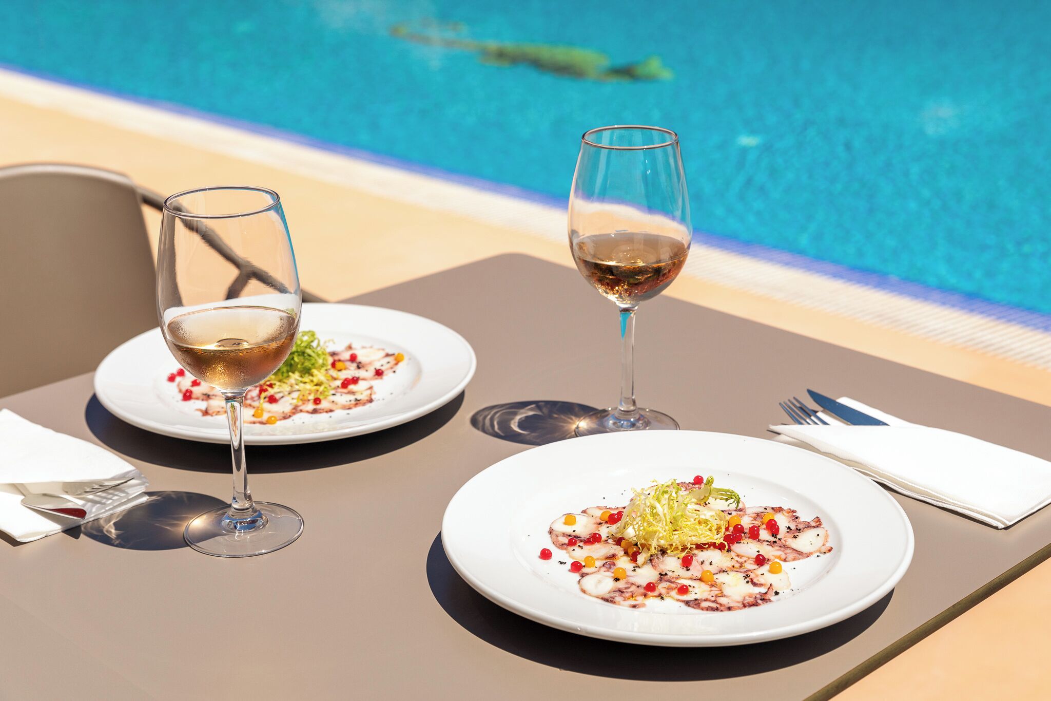 A dining table with two plates of food and two wine glasses, set against a poolside view.