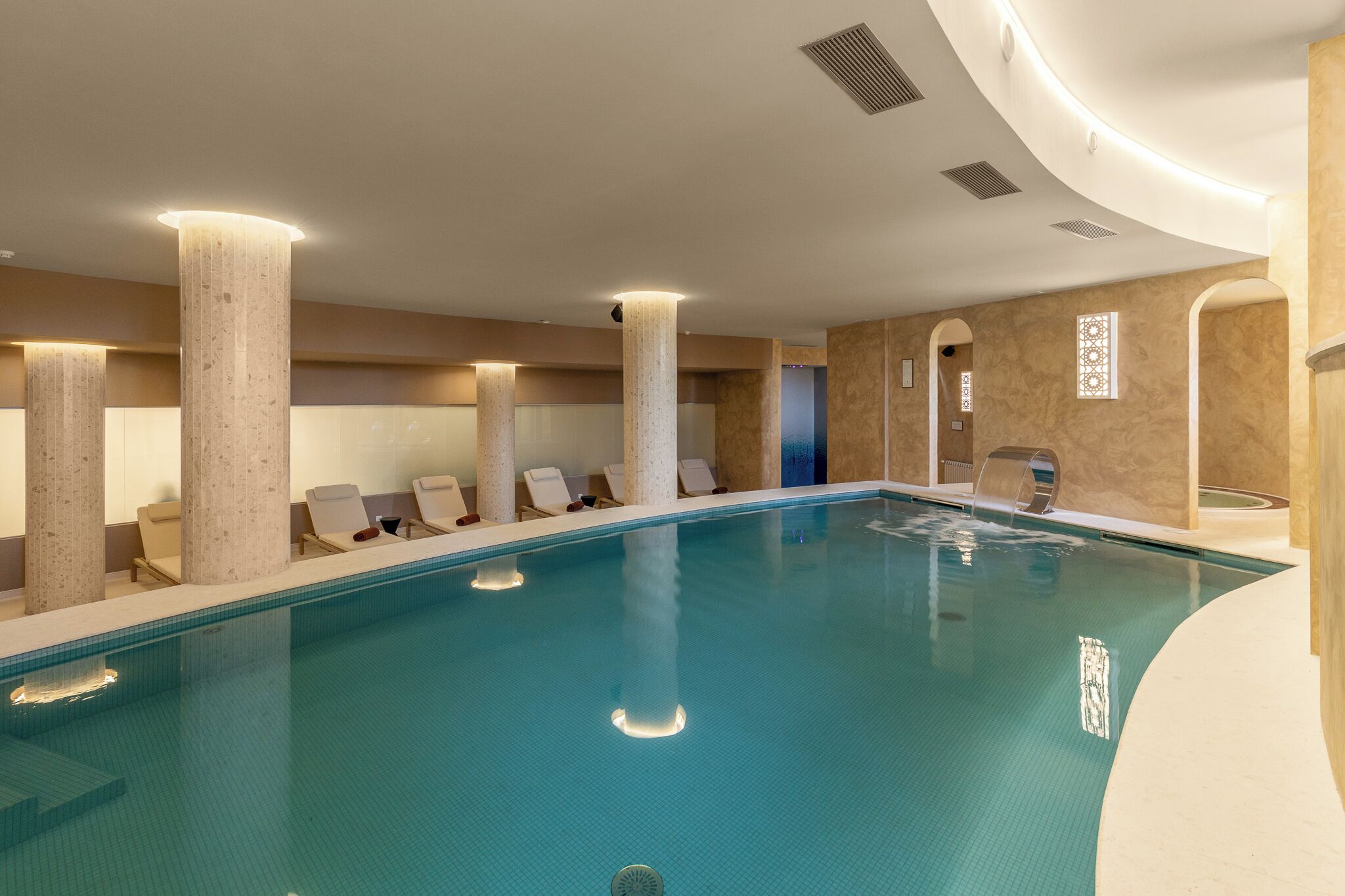 A swimming pool with lounge chairs and pillars, a curved ceiling, and a patterned wall.