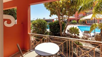 A balcony with a white table and wicker chairs, overlooking a swimming pool and lush greenery.