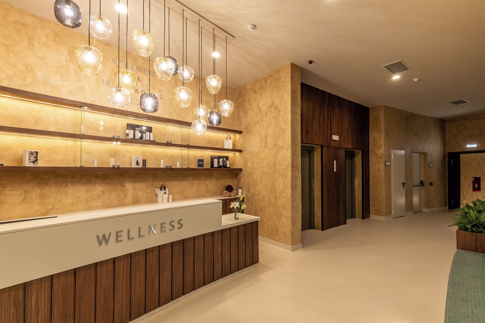 A reception area with a wooden reception desk labeled "WELLNESS," hanging crystal pendant lights, and a clean, modern interior design.