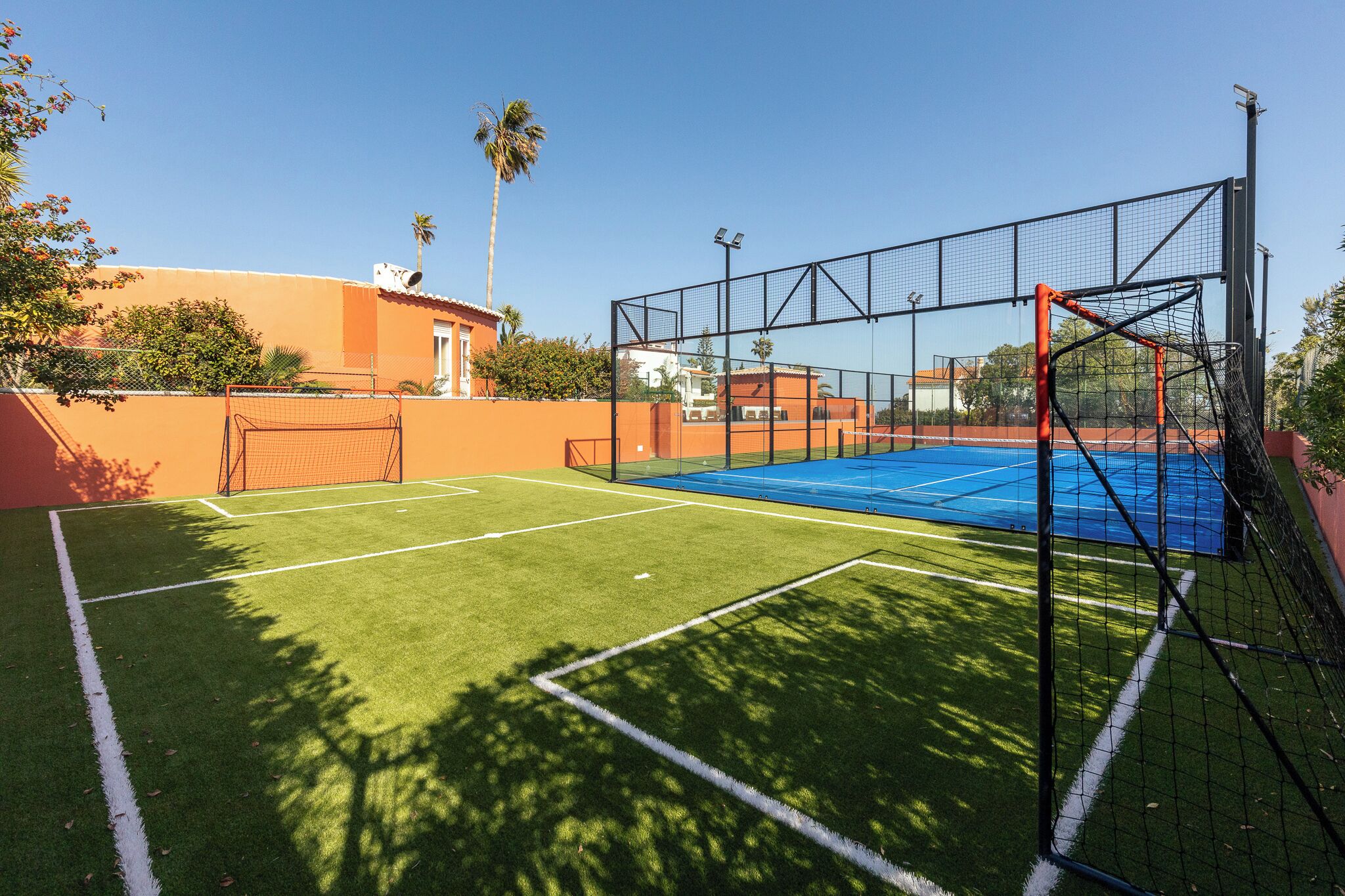 Un court de tennis extérieur, doté d’un filet et entouré d’une clôture, et situé à côté d’un bâtiment dont le toit est rouge.