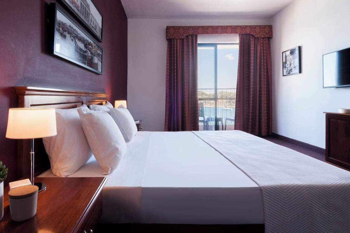 A hotel room with a large bed, a nightstand with a lamp, a TV, and a window with curtains.