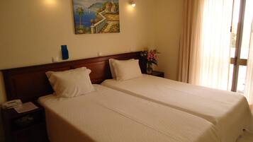 A hotel room with two beds, a nightstand with a phone, a painting of a coastal scene, and a window with curtains.