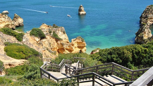 Paisaje costero con acantilados rocosos, mar azul cristalino y una escalera de madera que baja a la playa.