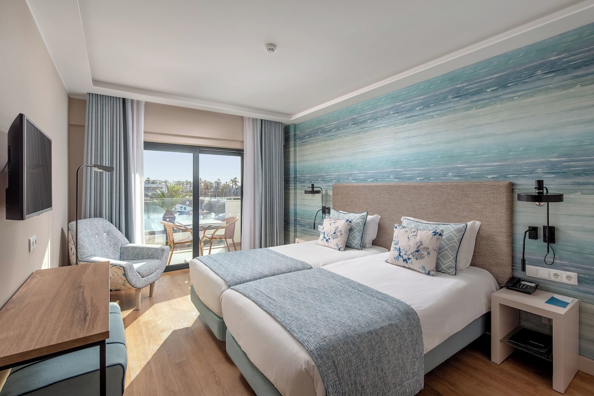 A modern hotel room with two beds, a dining area, and a view of the outdoors.